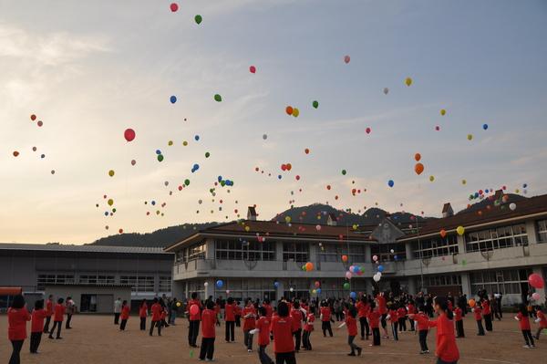 村雲小学校の運動場で、赤いティーシャツを着た沢山の人が色とりどり風船を空に飛ばしており、風船が上がっていくのを見上げている様子の写真